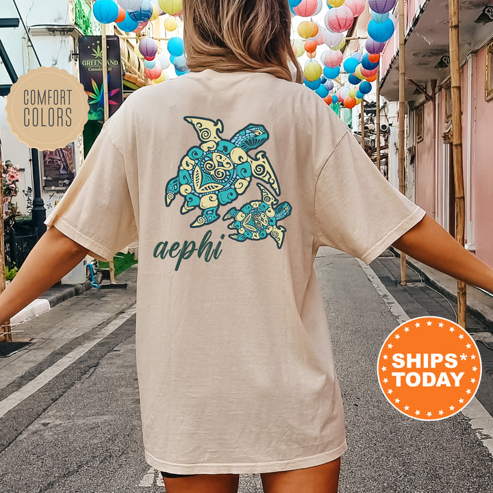A person wearing a beige t-shirt with a colorful turtle design and the text "aephi" printed on it, standing on a street with colorful lanterns hanging overhead.