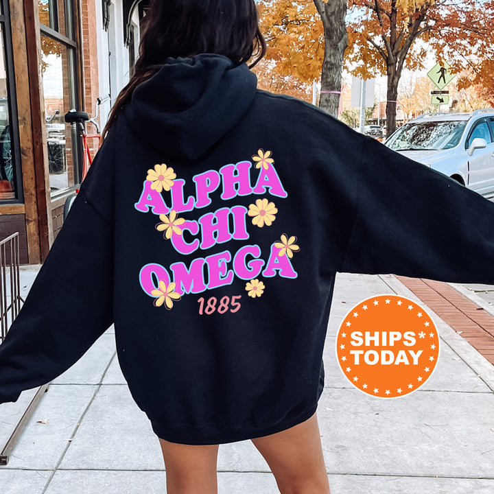 A person wearing a black hoodie with the text "ALPHA CHI OMEGA 1885" printed on it, standing on a sidewalk with a car and a tree in the background.