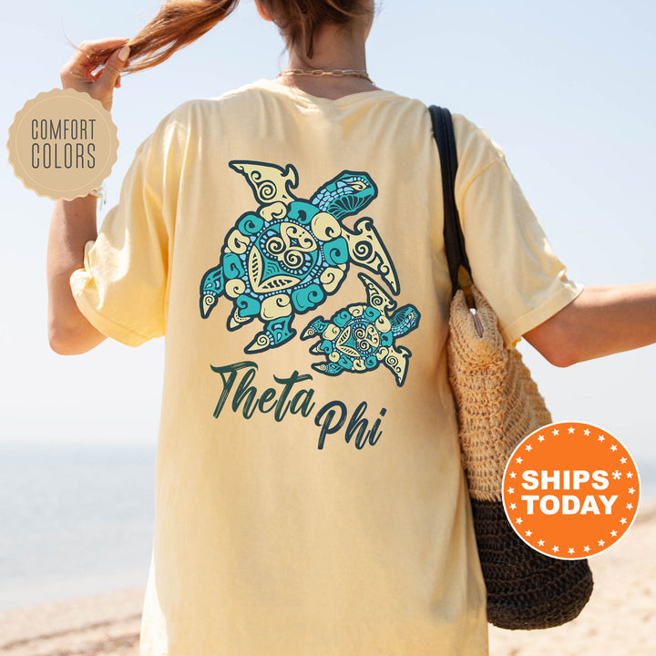 A person wearing a yellow t-shirt with a turtle design and the text "Theta Phi" printed on it, standing on a beach.
