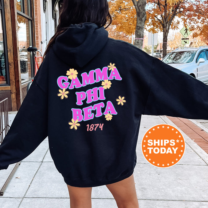 A person wearing a black hoodie with the text "Gamma Phi Beta" printed on it, standing on a sidewalk with a car and a building in the background.