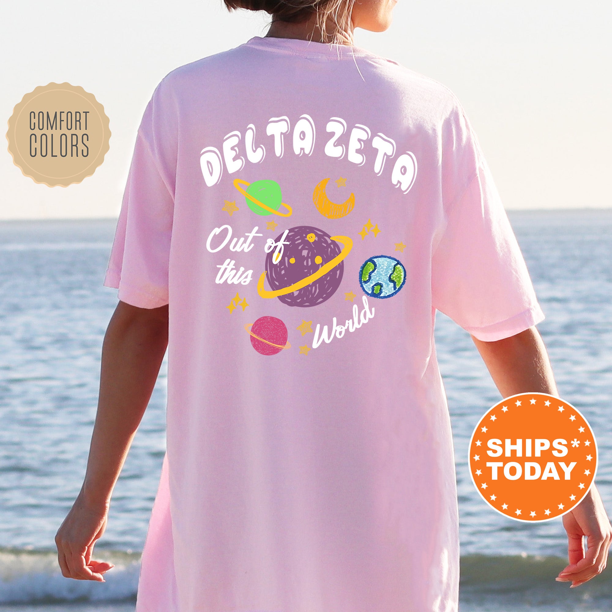 A person wearing a pink t-shirt with a graphic design of planets and stars, standing near the ocean.