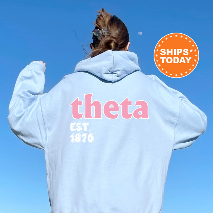 A person wearing a light blue hoodie with the text "theta EST. 1870" printed on it, standing against a clear blue sky.