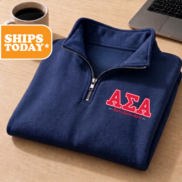 A navy blue zip-up jacket with the Greek letters 'ΑΕΛΑ' printed on it, along with the text 'ΑΕΛΑαση ΚΑΙΑΙΑ' below it. The jacket is placed on a wooden surface, and there is a cup of coffee and a laptop in the background.
