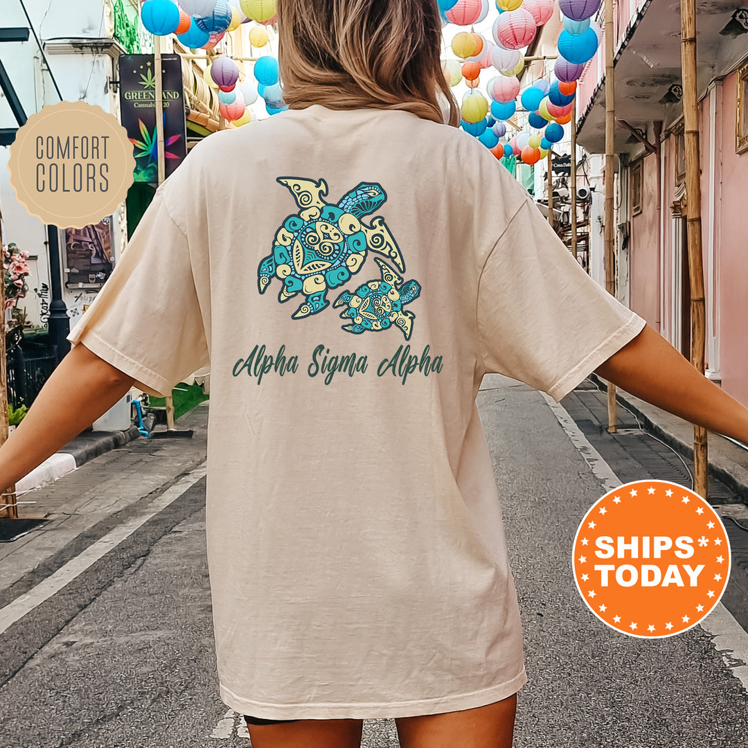 A person wearing a cream-colored t-shirt with a graphic of a turtle and the text "Alpha Sigma Alpha" printed on it, standing on a street with colorful lanterns hanging overhead.