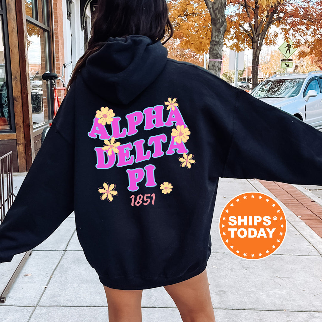 A person wearing a black hoodie with the text "ALPHA DELTA PI 1851" printed on it, standing on a sidewalk with a car and a building in the background.