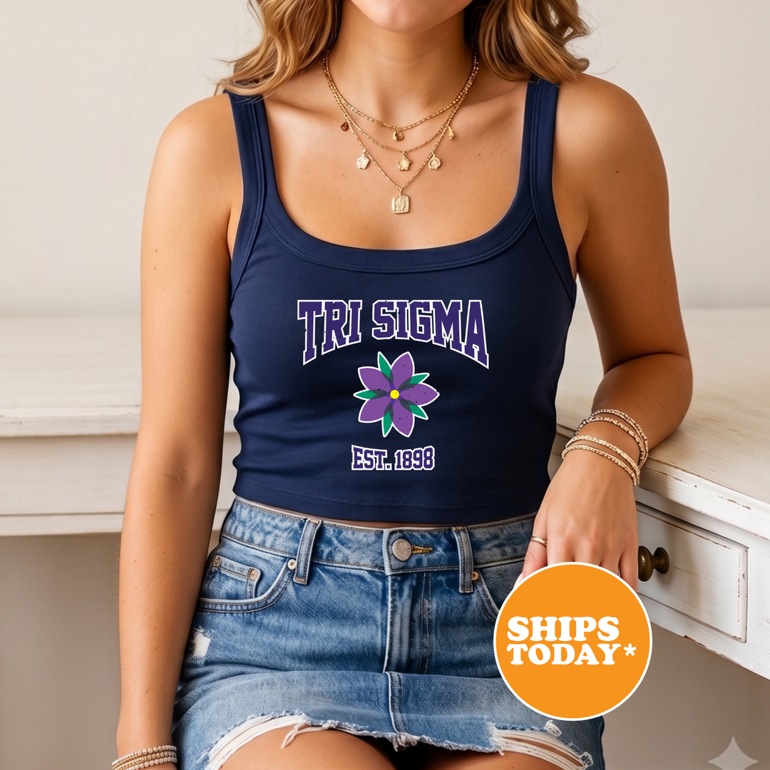 A woman wearing a navy blue crop top with the text "TRI SIGMA" and a purple flower graphic, paired with denim shorts. She is also wearing multiple necklaces and bracelets.
