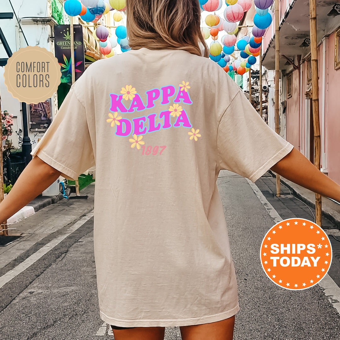 A person wearing a beige t-shirt with the text " Kappa Delta" printed on it, standing on a street with colorful lanterns hanging overhead.