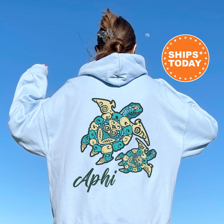 A person wearing a light blue hoodie with a colorful turtle design and the name 'Aphi' printed on it, standing against a clear blue sky.
