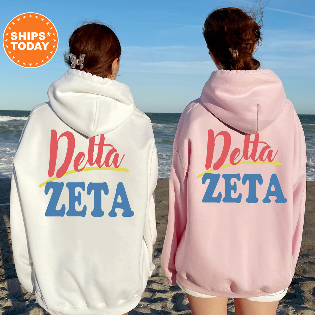 Two women standing on a beach, wearing matching white and pink hoodies with the text "Delta Zeta" printed on them.