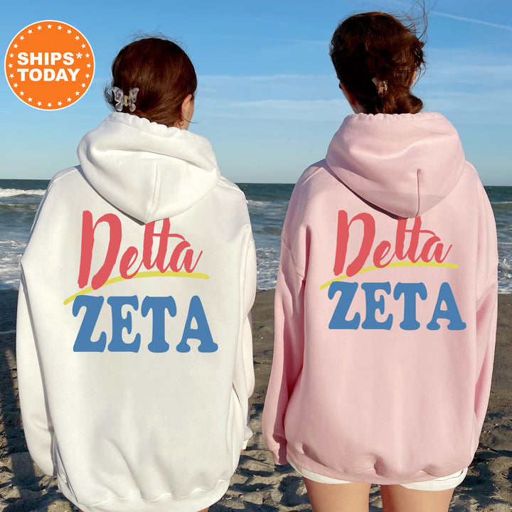 Two women standing on a beach, wearing matching white and pink hoodies with the text "Delta Zeta" printed on them.