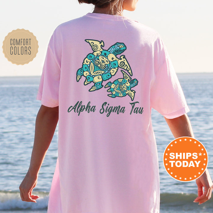 A person wearing a pink t-shirt with a graphic of a turtle and the text "Alpha Sigma Tau" printed on it, standing near the ocean.