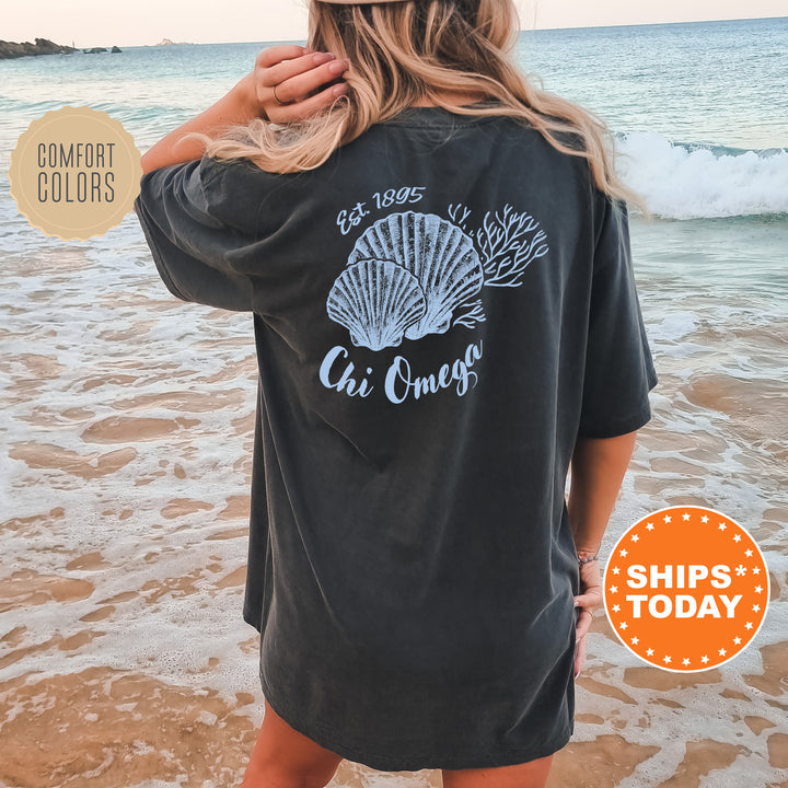 A woman wearing a black t-shirt with a graphic design of seashells and the text "Chi Omega" standing on a beach.