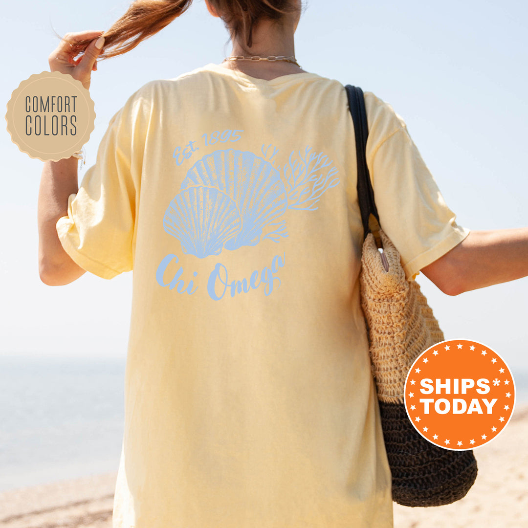 A woman wearing a yellow t-shirt with a blue design and text, standing on a beach with her hair in a ponytail.