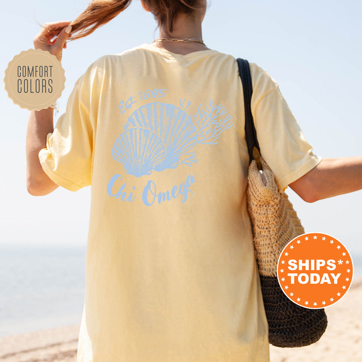A woman wearing a yellow t-shirt with a blue design and text, standing on a beach with her hair in a ponytail.