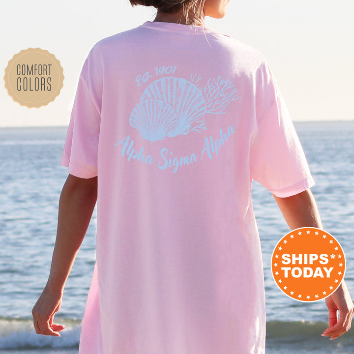 A person wearing a pink t-shirt with the text "Alpha Sigma Delta" printed on it, standing on a beach with the ocean in the background.