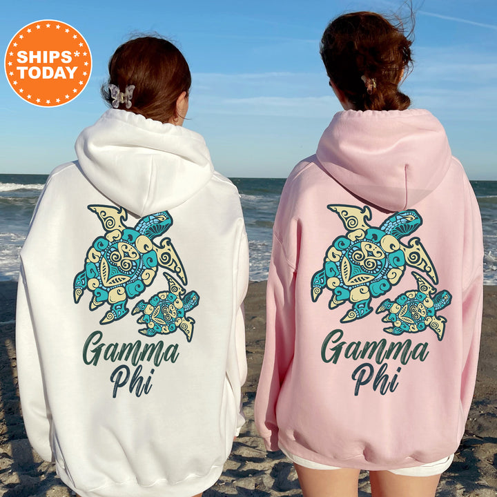 Two women wearing matching white and pink hoodies with a graphic of turtles on the back, standing on a beach with the ocean in the background.