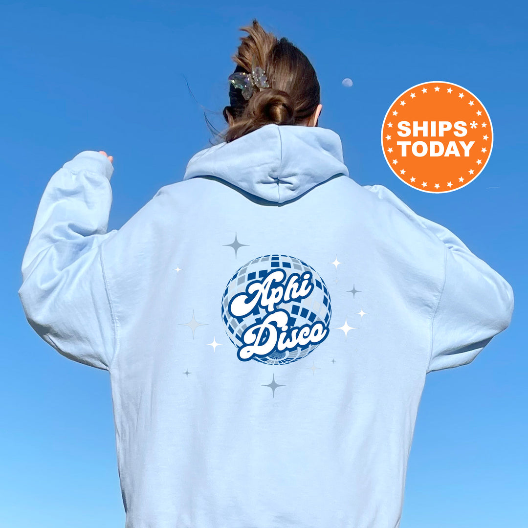 A person wearing a light blue hoodie with a graphic design on the back, including a disco ball and stars, is standing against a clear blue sky.