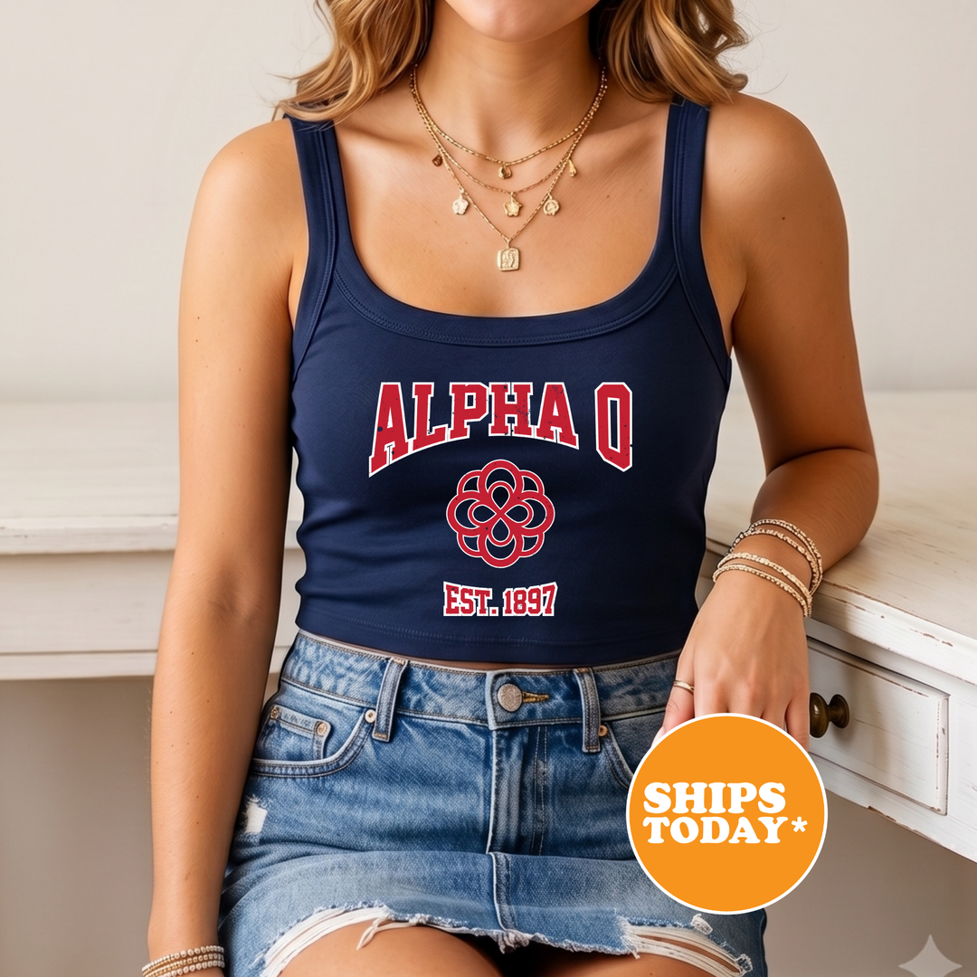 A woman wearing a navy blue crop top with the letters "ALPAQ" and a red design, paired with denim shorts, sits on a white surface. She is also wearing multiple necklaces and bracelets.