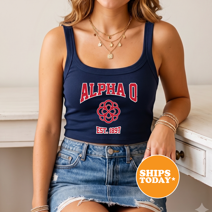 A woman wearing a navy blue crop top with the letters "ALPAQ" and a red design, paired with denim shorts, sits on a white surface. She is also wearing multiple necklaces and bracelets.
