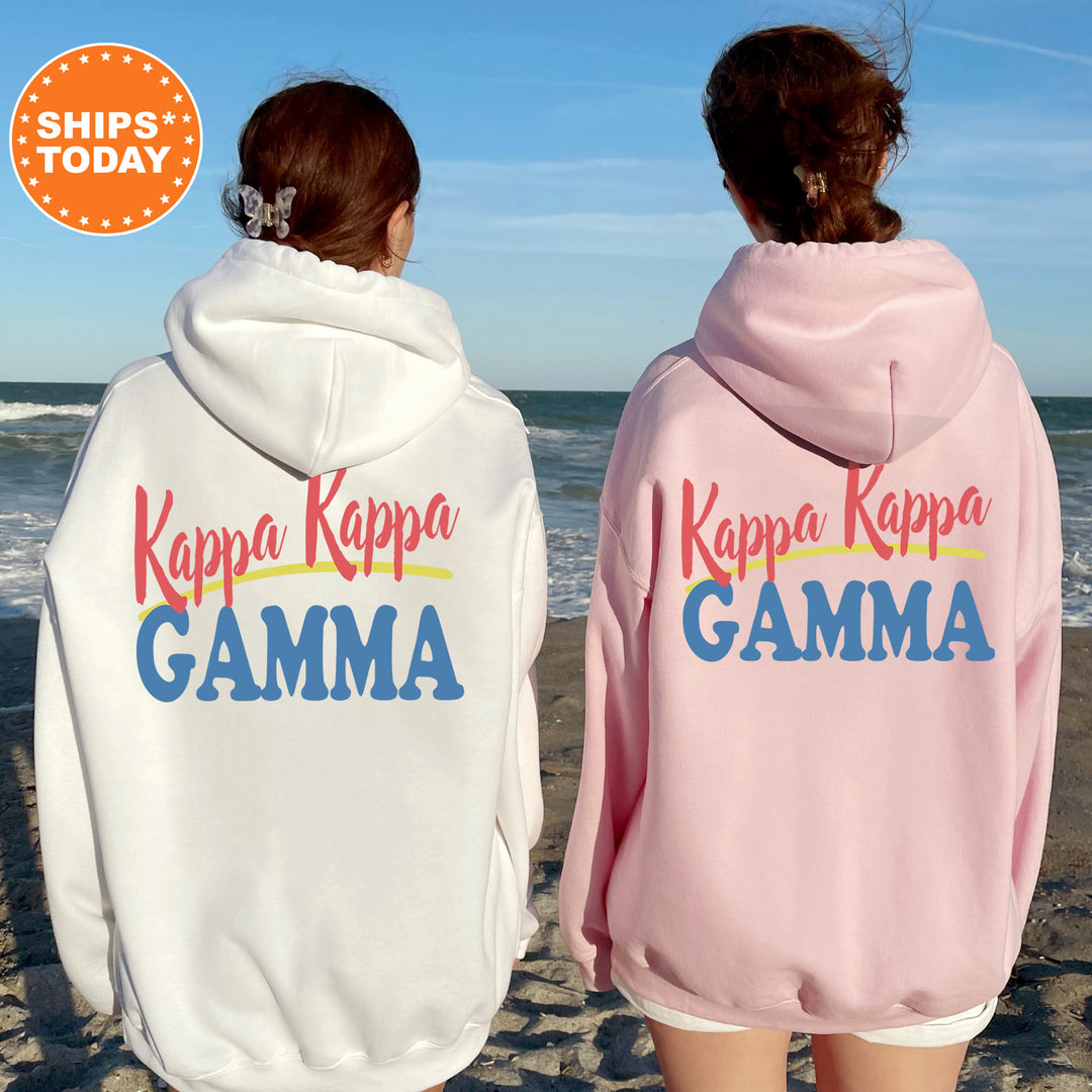 Two women wearing matching white and pink hoodies with the Kappa Kappa Gamma sorority logo on the back, standing on a beach with the ocean in the background.