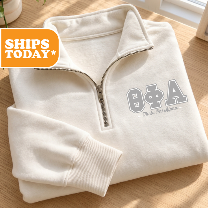 A white sweatshirt with the Greek letters "ΦΑΓ" printed on it, along with the text "Theta Phi Alpha" below the letters. The sweatshirt is folded and placed on a wooden surface, with a shipping label visible in the top left corner.