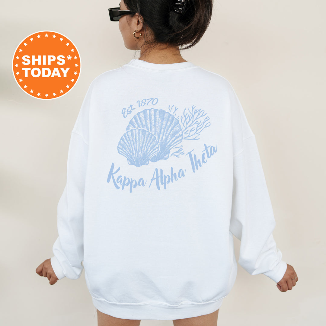 A woman wearing a white sweatshirt with a blue design featuring a shell and the text " Kappa Alpha Theta" printed on it.