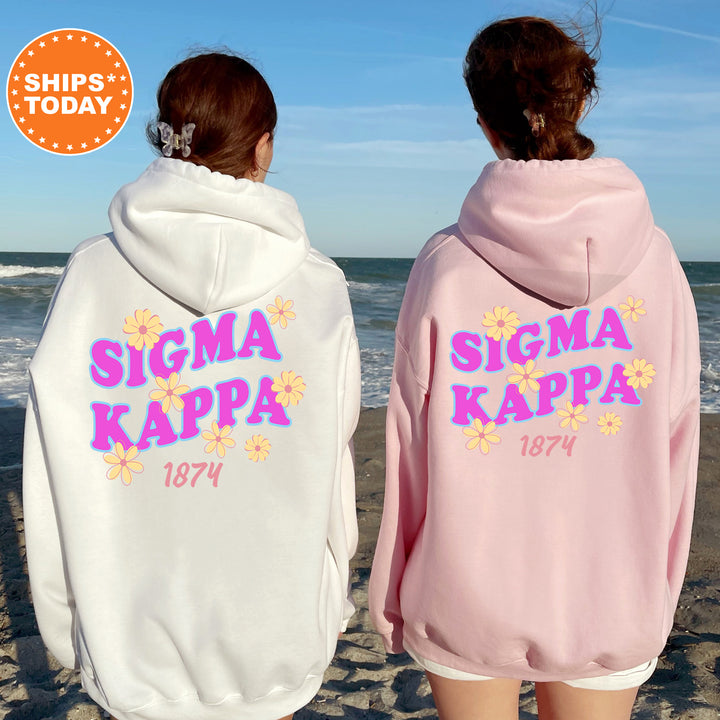 Two women wearing white and pink hooded sweatshirts with the Sigma Kappa logo and the year 1874 printed on them, standing on a beach with the ocean in the background.