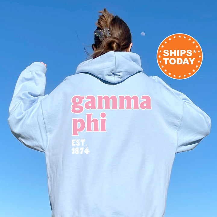 A person wearing a light blue hoodie with the text "gamma phi" and "EST. 1874" printed on it, standing against a clear blue sky.