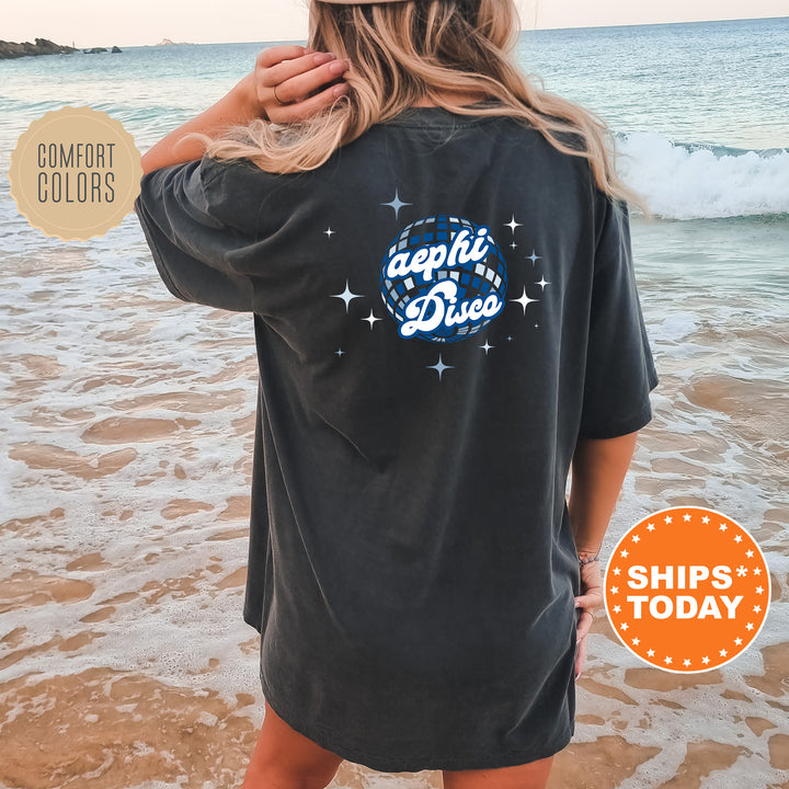 A woman wearing a black t-shirt with a graphic design on the back is standing on a beach, with the ocean and waves visible in the background.