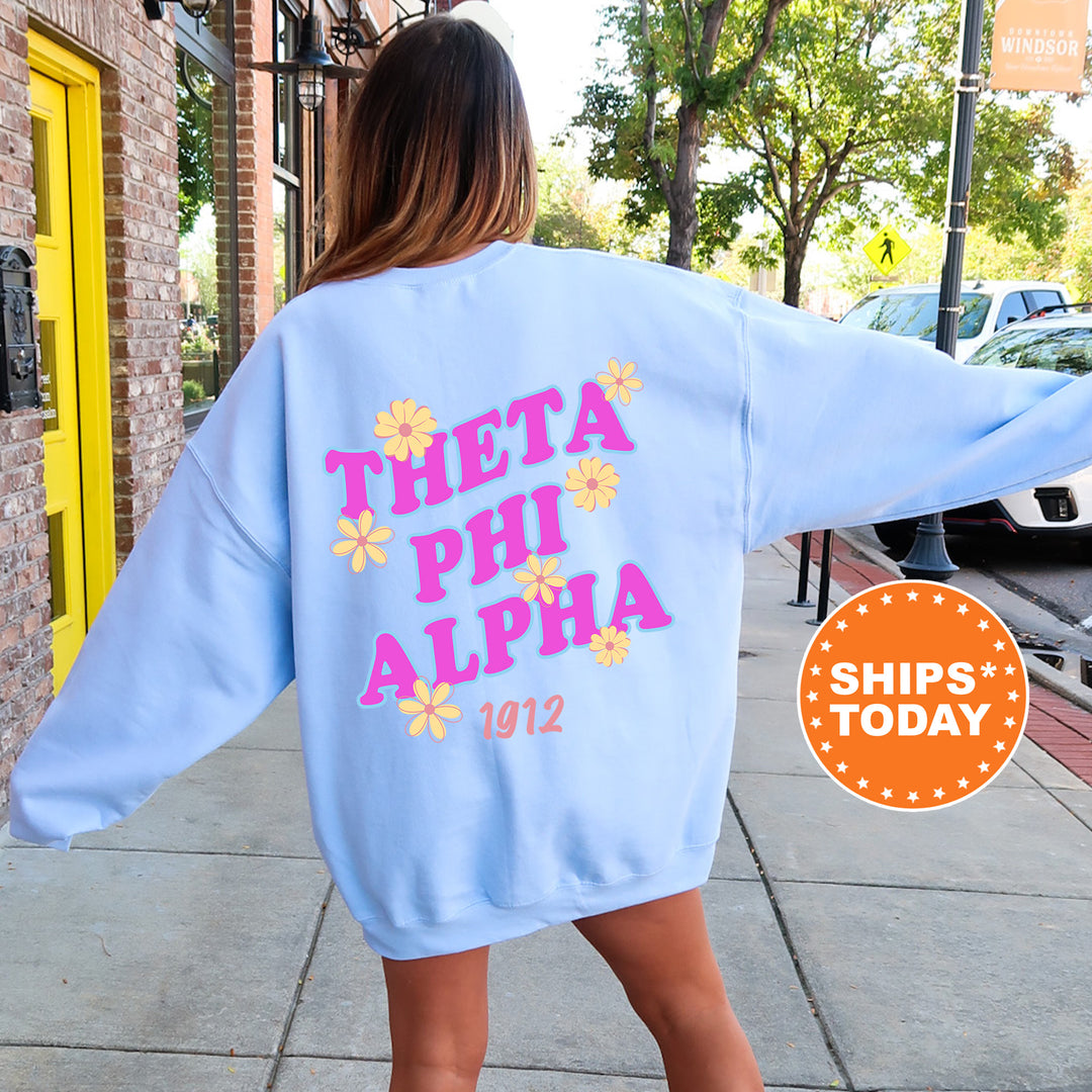 A woman wearing a light blue sweatshirt with the text "THETA PHI ALPHA 1912" printed on it, standing on a sidewalk.