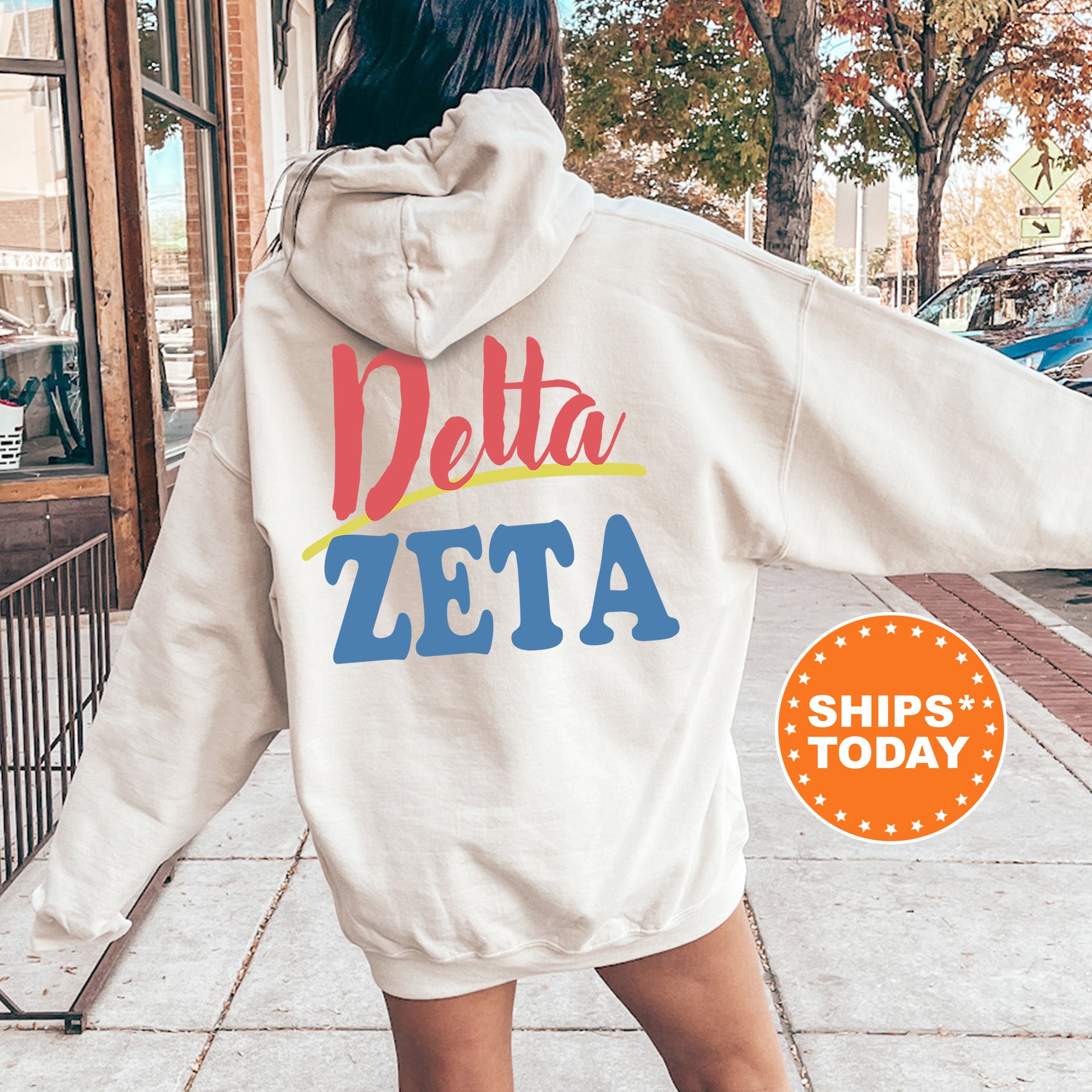 A person wearing a white hoodie with the Delta Zeta logo printed on it, standing on a sidewalk with a car and trees in the background.