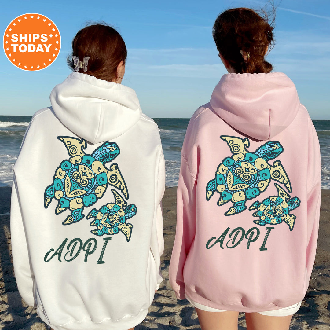Two women wearing hooded sweatshirts with a turtle design on the back, standing on a beach with the ocean in the background.