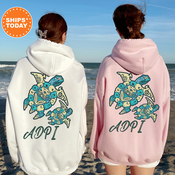 Two women wearing hooded sweatshirts with a turtle design on the back, standing on a beach with the ocean in the background.