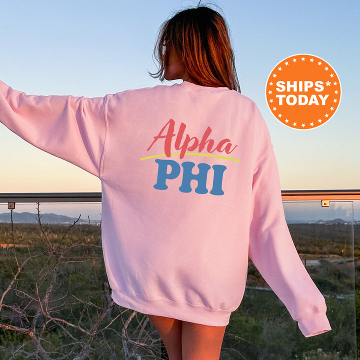 A woman wearing a pink sweatshirt with the text "Alpha Phi" printed on it, standing in front of a scenic outdoor landscape with mountains in the background.