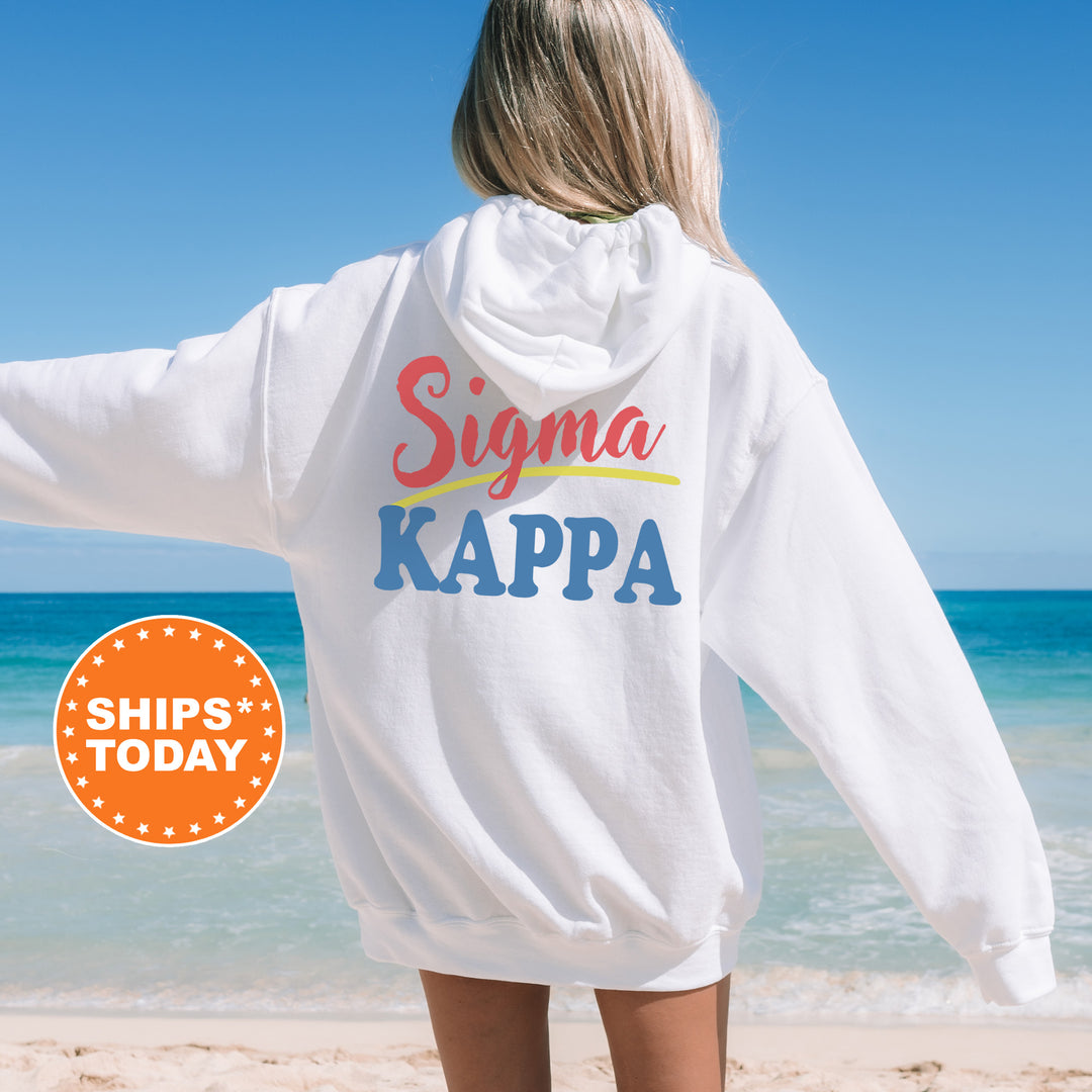 A woman wearing a white hoodie with the text "Signa Kappa" printed on it, standing on a beach with the ocean in the background.