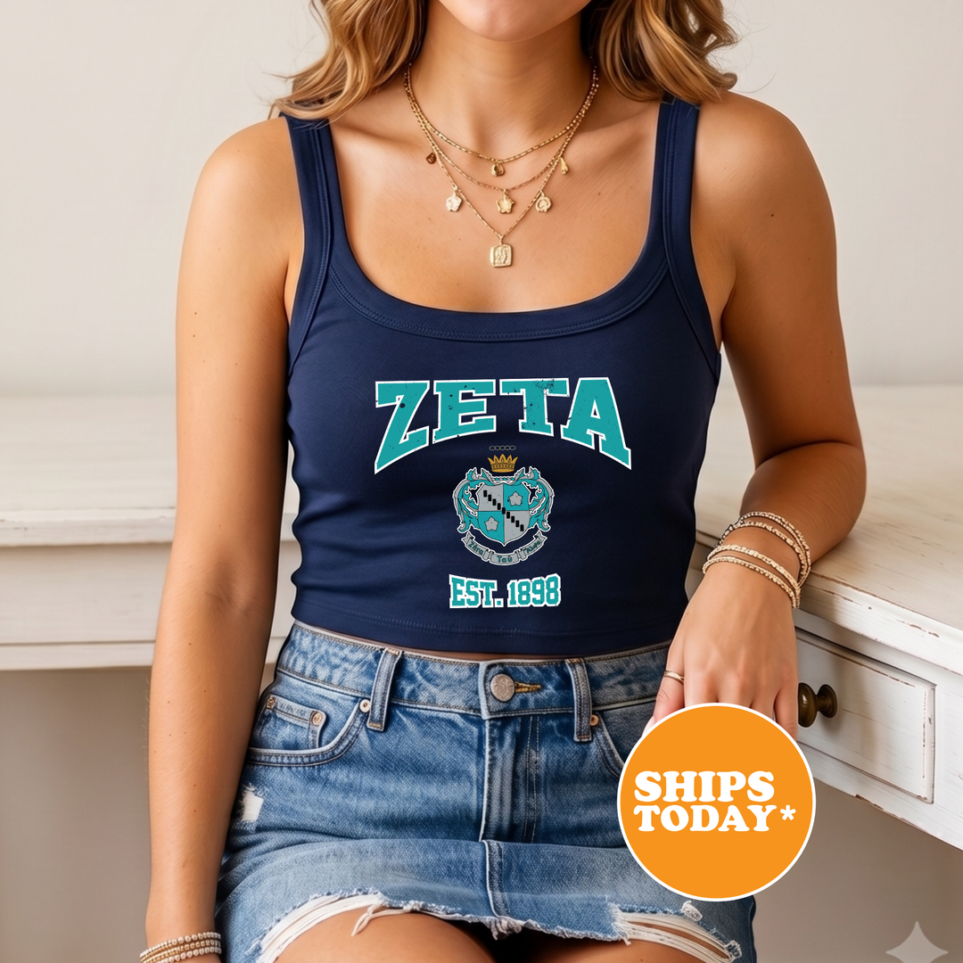 A woman wearing a navy blue crop top with the word "ZETA" printed on it, a denim skirt, and multiple necklaces. She is sitting on a white surface, and there is a circular orange sign with white text that says "SHIPS TODAY".
