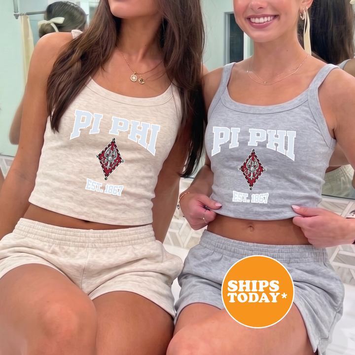 Two women wearing matching Pi Phi tank tops and shorts are posing together in front of a mirror.