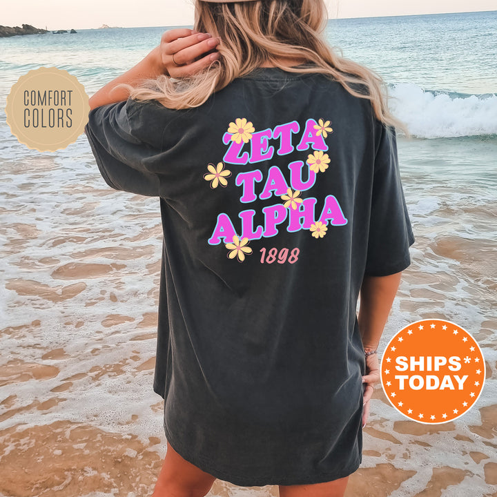 A woman wearing a black t-shirt with the text "ZETA Tau Alpha" printed on it, standing on a beach with waves in the background.