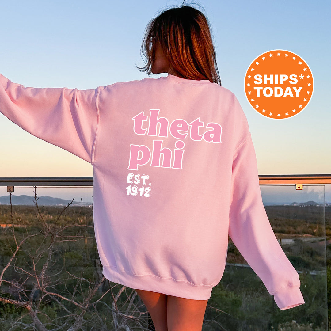 A woman wearing a pink sweatshirt with the text "theta phi" printed on it, standing in front of a scenic outdoor landscape.
