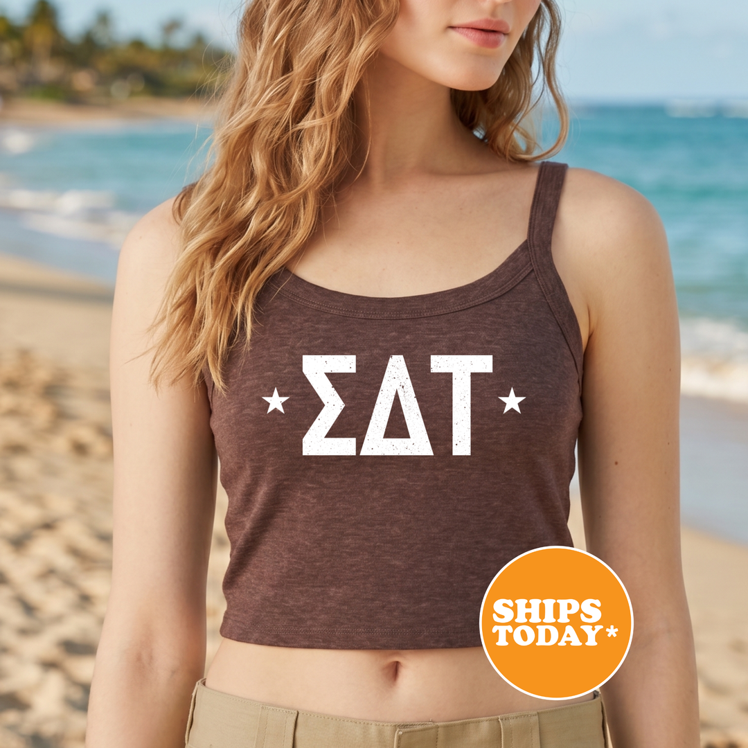 A woman wearing a brown tank top with the word "ΔΕΛΑ" printed on it, standing on a beach with the ocean in the background.