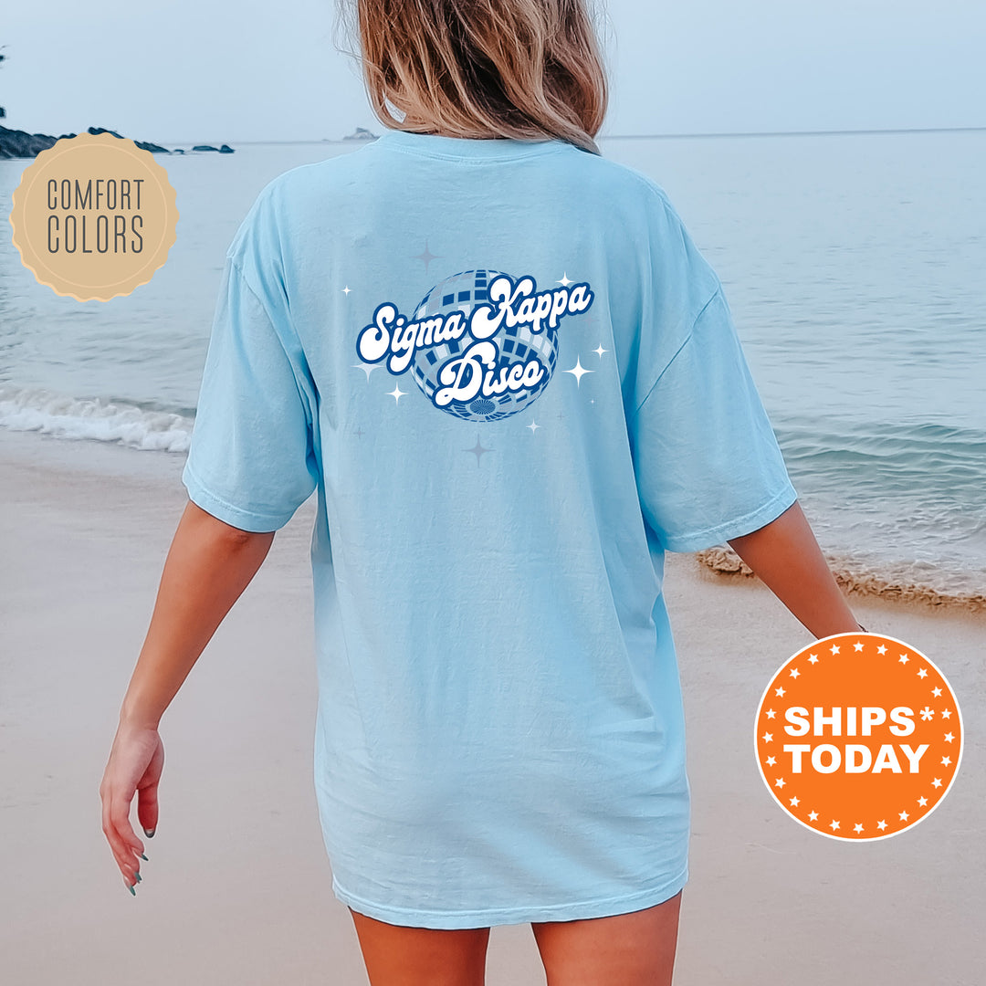 A woman wearing a light blue t-shirt with the Sigma Kappa Delta logo and text, standing on a beach with the ocean in the background.