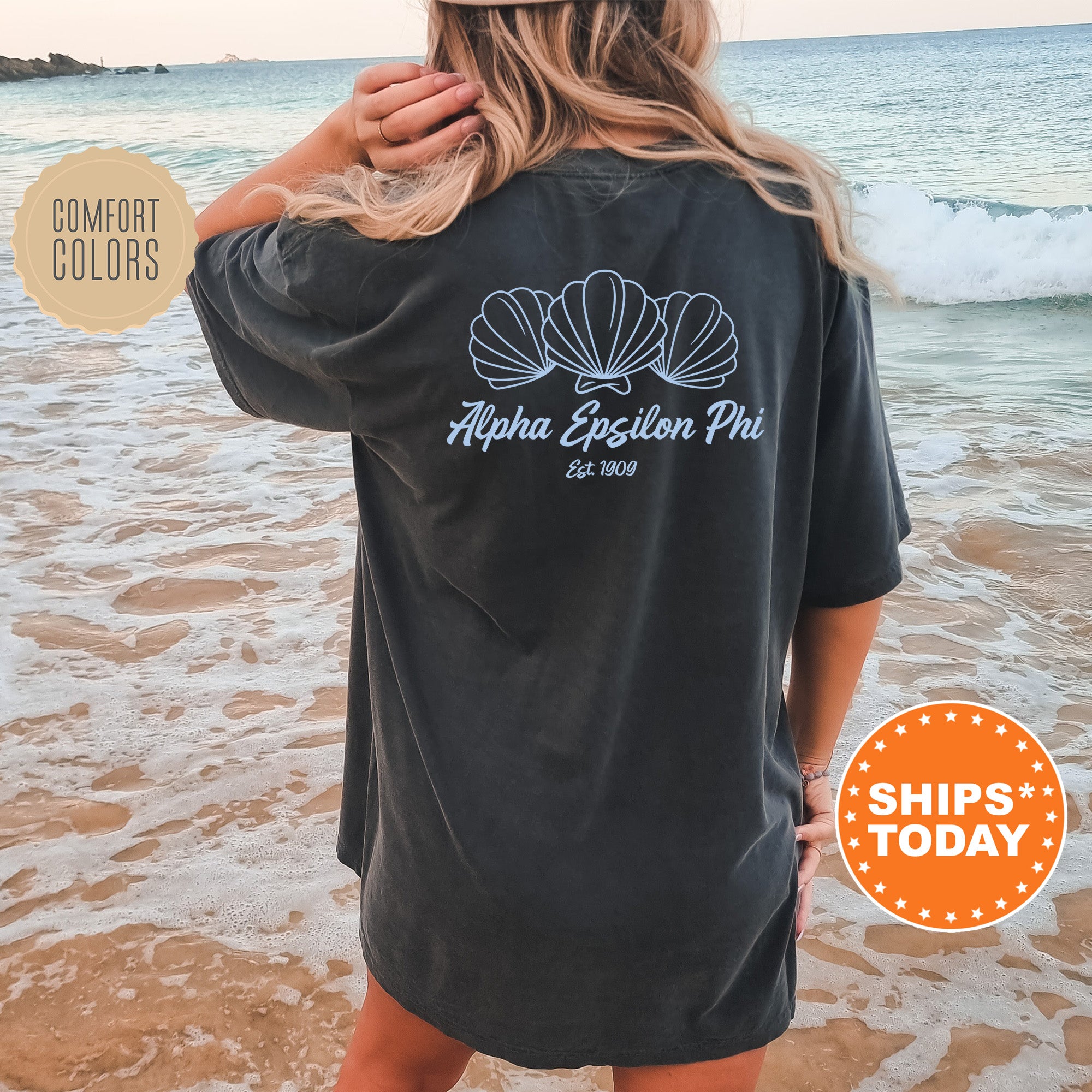 A woman wearing a black t-shirt with a graphic design of shells on the back, standing on a beach with the ocean in the background.
