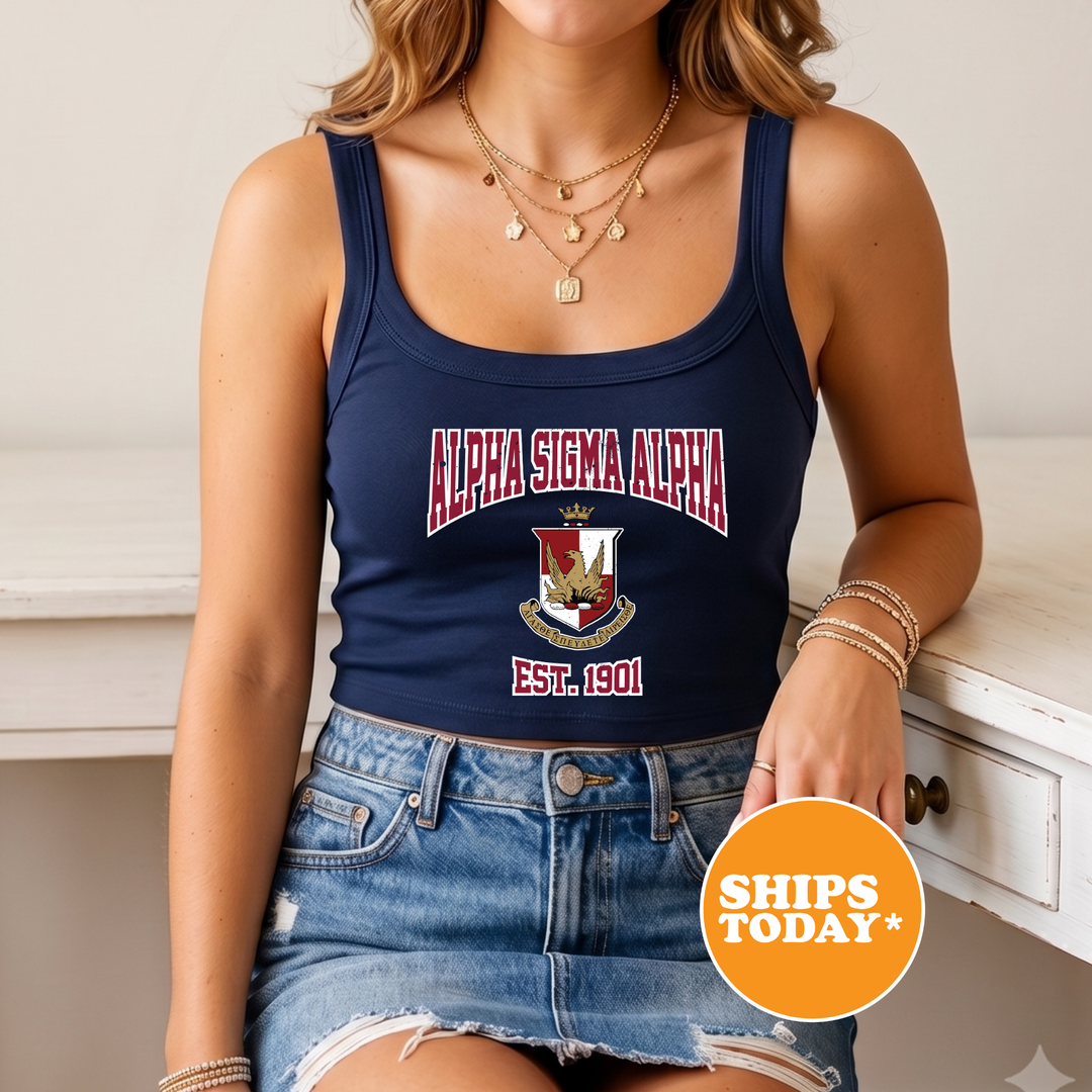 A woman wearing a navy blue tank top with the text "ALPHA SIGMA ALPHA EST. 1901" and a denim skirt, along with multiple necklaces. She is sitting on a white surface, and there is a "SHIPS TODAY" sign in the bottom right corner of the image.