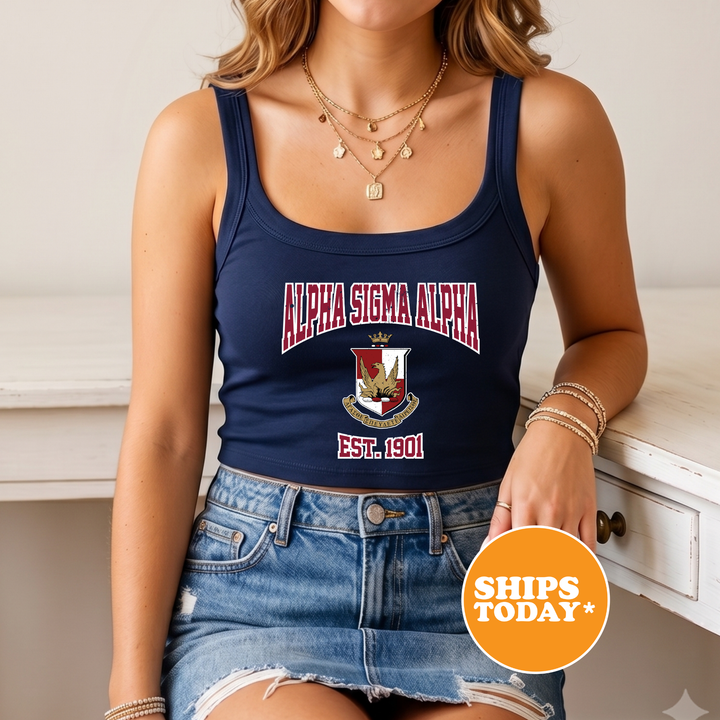 A woman wearing a navy blue tank top with the text "ALPHA SIGMA ALPHA EST. 1901" and a denim skirt, along with multiple necklaces. She is sitting on a white surface, and there is a "SHIPS TODAY" sign in the bottom right corner of the image.