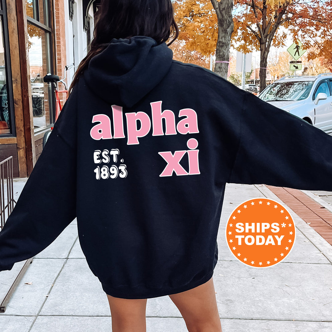 A person wearing a black hoodie with pink text and logos, standing on a sidewalk with a car and a building in the background.