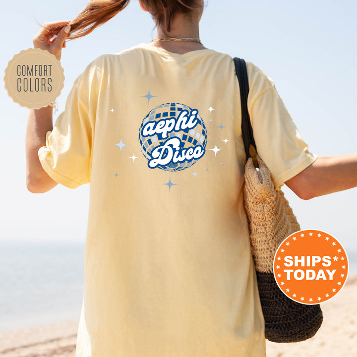 A person wearing a yellow t-shirt with a graphic design and text, standing on a beach with a handbag and a sign that says "SHIPS TODAY".