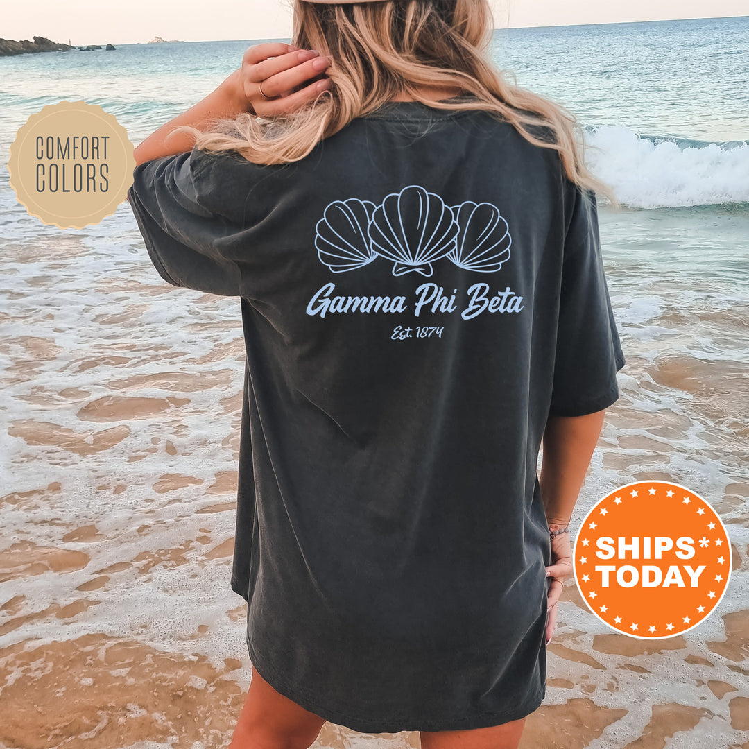 A woman wearing a black t-shirt with a graphic design of shells on the back, standing on a beach with the ocean in the background.