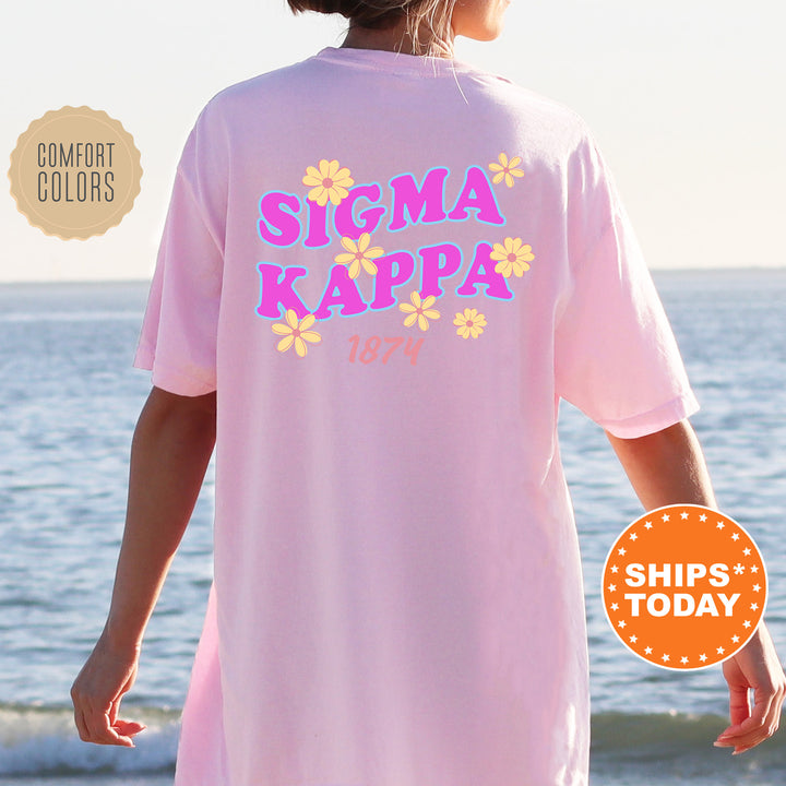 A person wearing a pink t-shirt with the text "SIGMA KAPPAS" and "1874" printed on it, standing near the ocean.