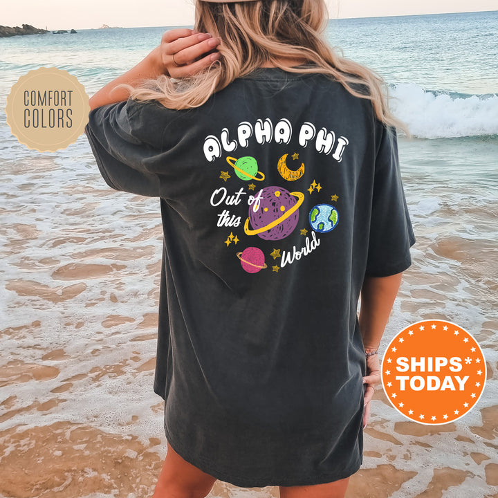 A woman wearing a black t-shirt with a graphic design of planets and stars on the back, standing on a beach with the ocean in the background.