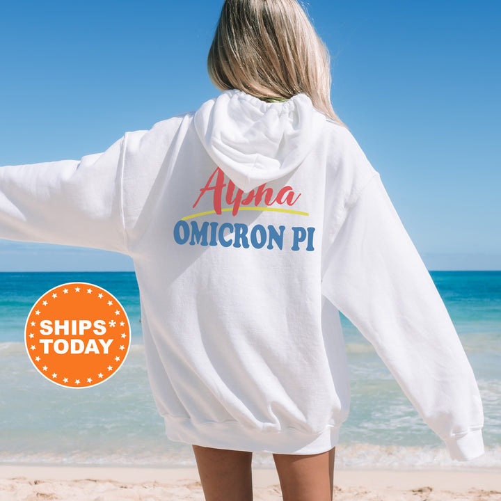 A person wearing a white hoodie with the text "Alpha Omega Pi" printed on it, standing on a beach with the ocean in the background.